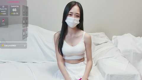 Media: Video of an Asian woman with long black hair, wearing a white mask, crop top, and pants, sitting on a bed with white sheets, in a minimalist bedroom.