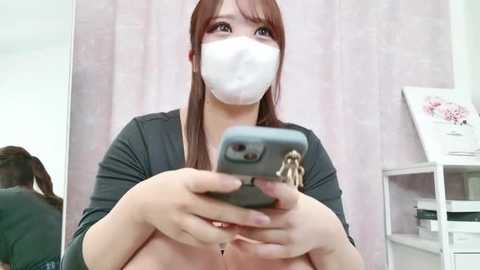 Media: Video of an Asian woman with light skin, long brown hair, wearing a white surgical mask and a black long-sleeved shirt, taking a selfie with a smartphone, in a room with light pink curtains and a white shelf with books.