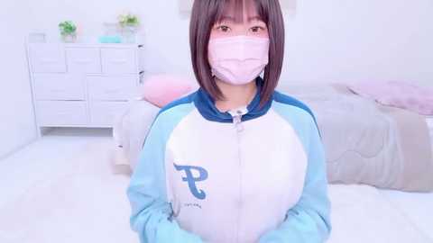 Media: Video of an Asian woman with straight, shoulder-length brown hair, wearing a light blue and white tracksuit with a \"P\" logo, a pink face mask, and a white face covering. She is in a bright, minimalist bedroom with white furniture.