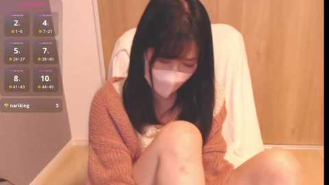 Media: A video of an Asian woman with black hair, wearing a beige mask, sitting on a wooden chair in a room with a white wall and wooden door. She's hunched over, appearing distressed.