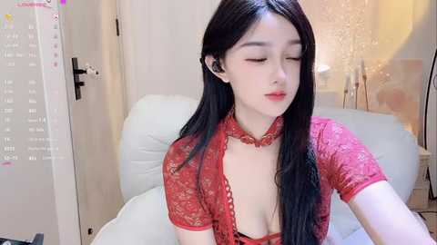 Media: Video of a young East Asian woman with long black hair, wearing a revealing red lace top, sitting on a white couch in a modern room with wooden accents.