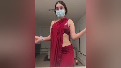 Media: Video of a young woman with light skin, dark hair, and glasses, wearing a maroon saree, mask, and scrunchie, in a modern, white-walled room.