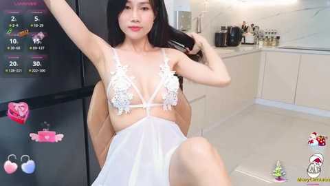 Media: Video of an Asian woman with long black hair, wearing a sheer white lingerie dress with floral lace details, sitting in a modern kitchen with white cabinets and a black refrigerator.