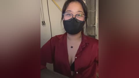 Media: Video of an Asian woman with glasses and black face mask, wearing a maroon button-up shirt with a black bra visible, standing in a dimly lit room with a yellow power cord in the background.