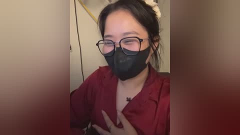 Media: Video of an Asian woman with fair skin, wearing black-rimmed glasses, a black face mask, and a red shirt, with a necklace, sitting indoors, hands to chest.