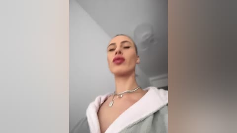 Media: Video of a fair-skinned, blonde woman with closed eyes, wearing a green robe, silver necklace, and red lipstick. She stands in a white room with a grey wall and a ceiling fan.