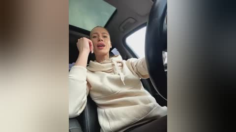 Media: A candid video of a woman with a light complexion, wearing a beige hoodie, driving a car, with a visible sun visor in the background.