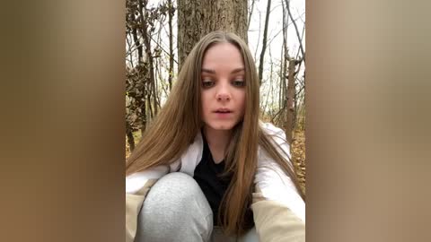 Media: A video of a young woman with long, straight brown hair, wearing a white jacket over a black top and grey sweatpants, kneeling in a forest.