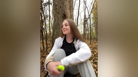 Media: Video of a young woman with long brown hair, wearing a white jacket over a black shirt, sitting on the forest floor, holding a neon green ball, surrounded by autumn leaves and tall trees.