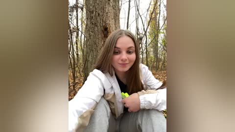 Media: A video of a young Caucasian woman with long, straight brown hair, sitting on the ground in a forest. She wears a white jacket over a black shirt, with gray pants. The background features tall trees and fallen leaves.