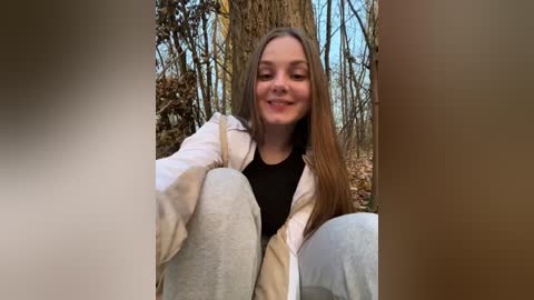 Media: Video of a smiling young woman with long brown hair, wearing a white jacket over a black shirt, seated on a bench in a forest with bare trees and autumn leaves.