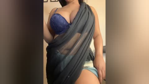 Media: Video of a light-skinned woman with a voluptuous figure wearing a blue lace bra and a dark gray fabric draped over her shoulder, exposing her right breast. She is also wearing light blue panties. The background is a neutral-toned wall with framed pictures.