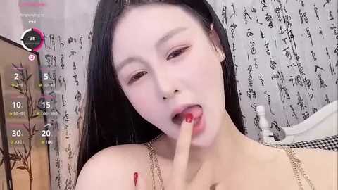 Media: Video of an Asian woman with long black hair and fair skin, wearing a gold chain necklace, licking her finger, in a room with Japanese kanji wallpaper.