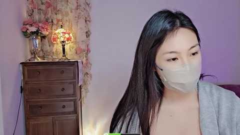 Media: Video of a young East Asian woman with long black hair, wearing a gray cardigan and a face mask, standing indoors near a vintage wooden dresser with a floral lamp and vase.