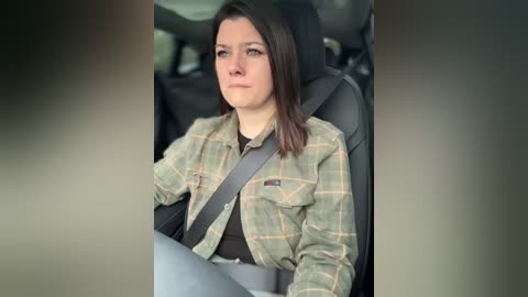 Video of a young woman with shoulder-length dark hair, wearing a plaid shirt and gray seatbelt, seated in a car, looking thoughtful.