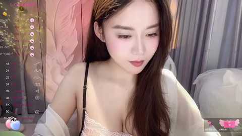 Media: Video of a young Asian woman with fair skin, long brown hair, wearing a pink lace bra, sitting indoors with a soft-focus background.