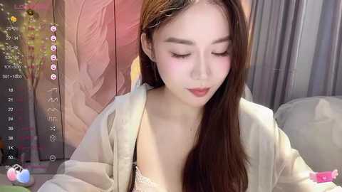 Media: Video of a young East Asian woman with long brown hair, light skin, and closed eyes, wearing a light-colored robe, sitting on a couch, with a digital camera screen in the background displaying her face and time.