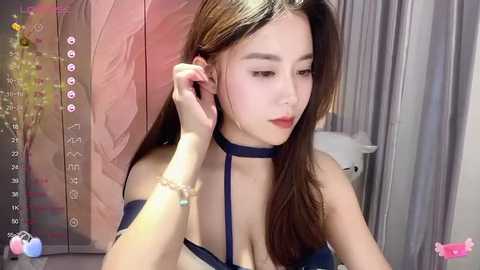 Media: A video of an East Asian woman with long brown hair and fair skin, wearing a blue choker and bra, adjusting her hair in a dimly lit room with floral wallpaper.