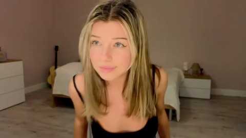 Media: A video of a blonde woman with shoulder-length hair, wearing a black tank top, leaning slightly forward in a beige room with light wooden floors and a bed with white sheets.