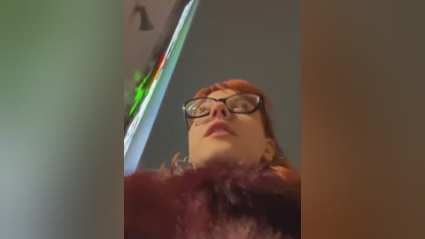 Media: A video of a young woman with short red hair and glasses, wearing a purple fur-trimmed coat, looking upward at a green and red neon sign, blurred background.