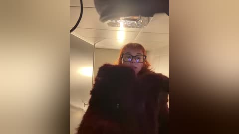 Media: Video of a woman with red hair, wearing glasses and a dark coat, standing in a dimly lit, narrow hallway with a fluorescent light above.