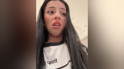 Media: Video of a young woman with long black hair, wearing a white \"Spirit\" t-shirt, with a neutral expression, blurred background.