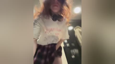 Media: Video of a young woman with curly red hair, wearing a white t-shirt and plaid skirt, in a dimly lit room, blurry background, leaning forward with a playful expression.