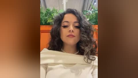 Media: Video of a young woman with curly dark hair, wearing a white top and pearl necklace, seated against a backdrop of green plants and wooden paneling.