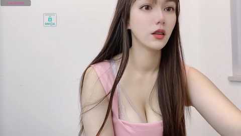 Media: Video of a pale-skinned, young East Asian woman with long, straight dark brown hair, wearing a pink tank top, looking contemplative against a white tiled wall with a green sign.