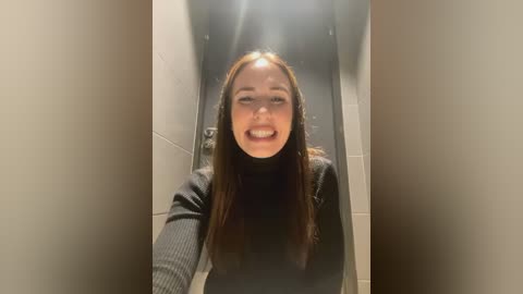 Media: Video of a young woman with long brown hair, smiling, wearing a black turtleneck sweater, in a narrow, dimly lit hallway with beige walls and a bright light source above her head.