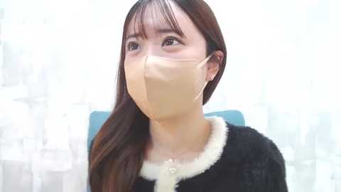 Media: Video of an Asian woman with long brown hair, wearing a black fur-trimmed cardigan, beige mask, and looking up thoughtfully. Background features white and blue abstract patterns.