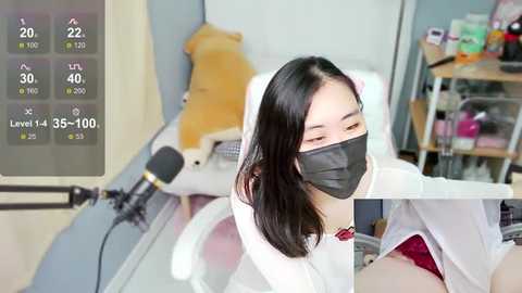 Media: Video of an East Asian woman with shoulder-length black hair, wearing a white lab coat and black face mask, sitting at a desk, surrounded by medical equipment, including a stethoscope and a thermometer display.