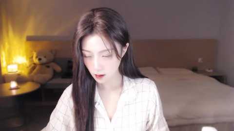 Media: A video of a young Asian woman with long, straight black hair, wearing a white plaid pajama top, standing in a dimly lit bedroom with a wooden headboard and a teddy bear on a nightstand.