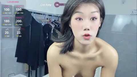 Media: Video of a young, slender East Asian woman with fair skin and dark hair, looking directly at the camera. She has a neutral expression, wearing only a white towel. Background features a gray tiled wall and a black coat hanging on a hook.