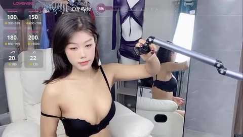 Media: A video of an Asian woman with long black hair, wearing a black bra, posing in a modern, minimalist room with a mirror and hanging clothes.