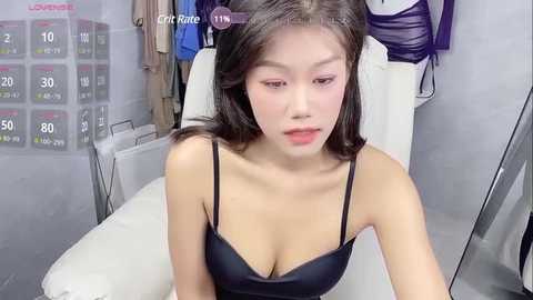 Media: Video of a young Asian woman with long dark hair in a black camisole, sitting on a white chair in a minimalistic room.