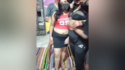 Media: Video of three people in a casual indoor setting. A woman in a red crop top, black skirt, and face mask stands center, flanked by two others. Background includes a colorful striped rug and a partially visible wall.