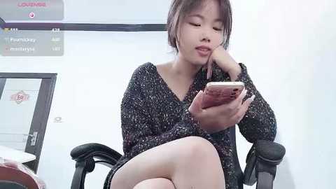 Media: Video of an Asian woman with short brown hair, wearing a black knit sweater, seated in a black office chair, holding a smartphone, in a brightly lit office with white walls and signage.