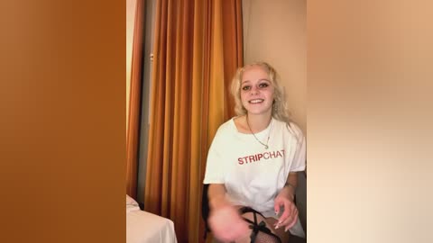 Media: A video of a blonde woman with long hair, wearing a white \"STRIPCHAT\" t-shirt, smiling, sitting in a room with beige walls and orange curtains.