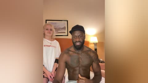 Media: Video of a shirtless, muscular Black man with a beard, sitting on a bed, next to a smiling, blonde, slim Caucasian woman in a white T-shirt. The room has a warm, beige tone, with a framed landscape painting on the wall.