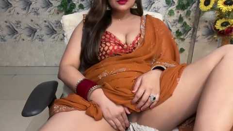 Media: Video of a voluptuous South Asian woman with fair skin, long brown hair, and red lipstick, wearing a brown sari with a red floral blouse, seated on a black office chair in a room with floral wallpaper and a sunflower bouquet.