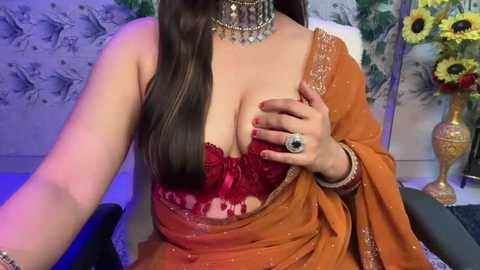 Media: Video of a fair-skinned woman with long dark hair, wearing an orange sari, a red lace bra, and gold jewelry, seated on a chair in a room with floral wallpaper and a vase of sunflowers.