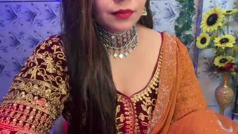 Media: Video of a woman with long brown hair, wearing a traditional Indian outfit with intricate gold embroidery on a deep maroon blouse, and an orange dupatta. She sports a silver choker with dangling pendants. Background includes sunflowers and floral wallpaper.