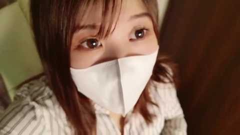 Media: A close-up video of an Asian woman with straight, brown hair, wearing a white surgical mask, brown eyes, and a white and gray striped shirt, set against a blurred background.