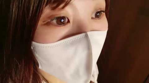 Media: Video of an Asian woman with short, dark hair, wearing a white surgical mask covering her nose and mouth, eyes looking directly at the camera.