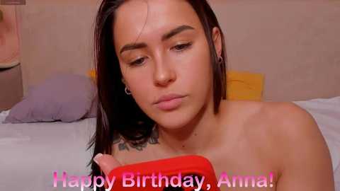 Media: A video of a young, light-skinned woman with dark hair, wearing a red shirt, sitting on a bed, looking sad. Text says \"Happy Birthday, Anna!\