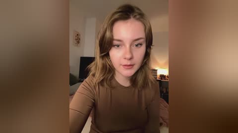 Media: A video of a young, light-skinned woman with shoulder-length, wavy, brown hair, wearing a brown top, sitting indoors. Background shows a dimly lit room with a lamp and a framed picture.