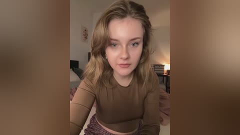 Media: A video of a young, fair-skinned woman with shoulder-length blonde hair, wearing a brown top and pink skirt, sitting on a carpeted floor in a dimly lit room with a lit lamp and minimal decor.