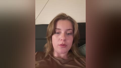 Media: A video of a young Caucasian woman with fair skin and shoulder-length blonde hair, wearing a brown shirt, looking introspective in a dimly lit room with neutral-colored walls.