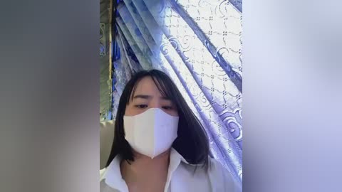 Media: Video of a young Asian woman with straight black hair, wearing a white surgical mask and a white shirt, standing in a room with a patterned blue and white curtain and a white wall.